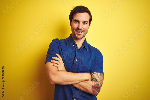 Young handsome man with tattoo wearing casual shirt standing over isolated yellow background happy face smiling with crossed arms looking at the camera. Positive person.