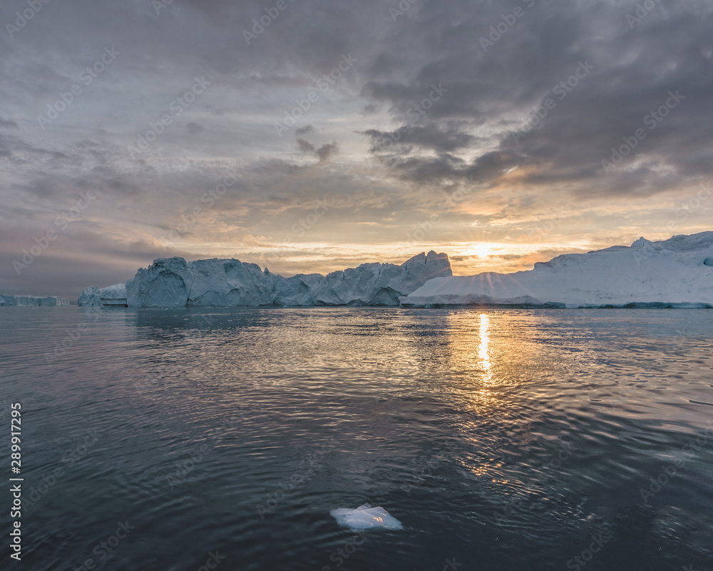 Arctic nature landscape with icebergs in Greenland icefjord with midnight sun sunset / sunrise in the horizon. Early morning summer alpenglow during midnight season. Ilulissat, West Greenland.