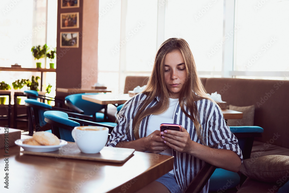 Young girl drinking morning cappuccino in cafe and and looking at the phone.