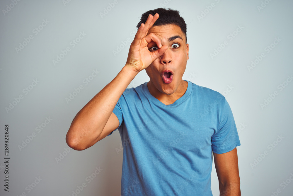 Young brazilian man wearing blue t-shirt standing over isolated white background doing ok gesture shocked with surprised face, eye looking through fingers. Unbelieving expression. Young brazilian man wearing blue t-shirt standing over isolated white background doing ok gesture shocked with surprised face, eye looking through fingers. Unbelieving expression.