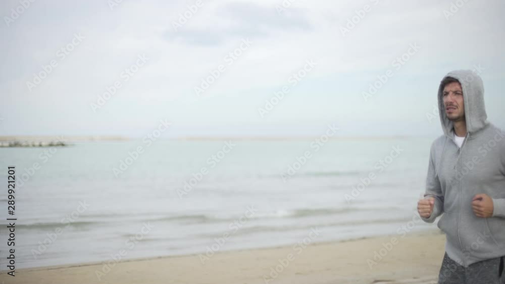 Young handsome man jogging running on the beach near the sea ocean. Slow motion of attractive guy with beard and a hoodie doing sport. Young handsome man jogging running on the beach near the sea ocean. Slow motion of attractive guy with beard and a hoodie doing sport.