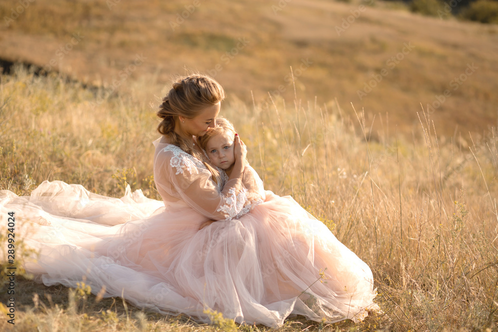 Young mother with a little daughter in pink dresses are sitting in the field. Mom hugs daughter, hugging her