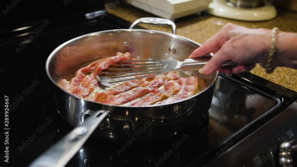 Bacon frying in stove top pan. Female chef turning bacon strips in silver skillet. Bacon frying in stove top pan. Female chef turning bacon strips in silver skillet.
