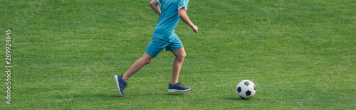 panoramic shit of kid in sportswear running on green grass with football