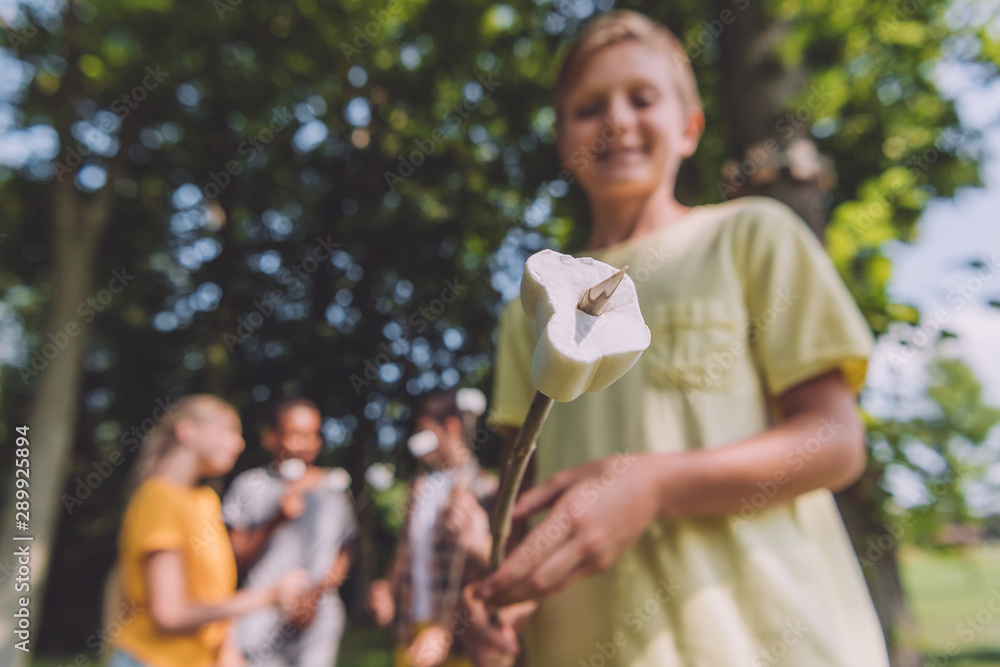 selective focus of happy boy holding sweet marshmallow on stick near friends selective focus of happy boy holding sweet marshmallow on stick near friends
