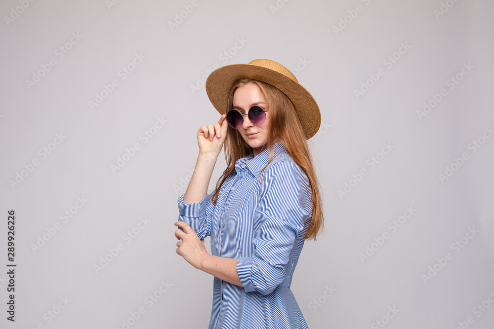 Side view of stylish female model wearing dress, glasses and straw hat posing on grey isolated background. Young blonde looking at camera while standing in studio. Concept of summer outfit. Side view of stylish female model wearing dress, glasses and straw hat posing on grey isolated background. Young blonde looking at camera while standing in studio. Concept of summer outfit.