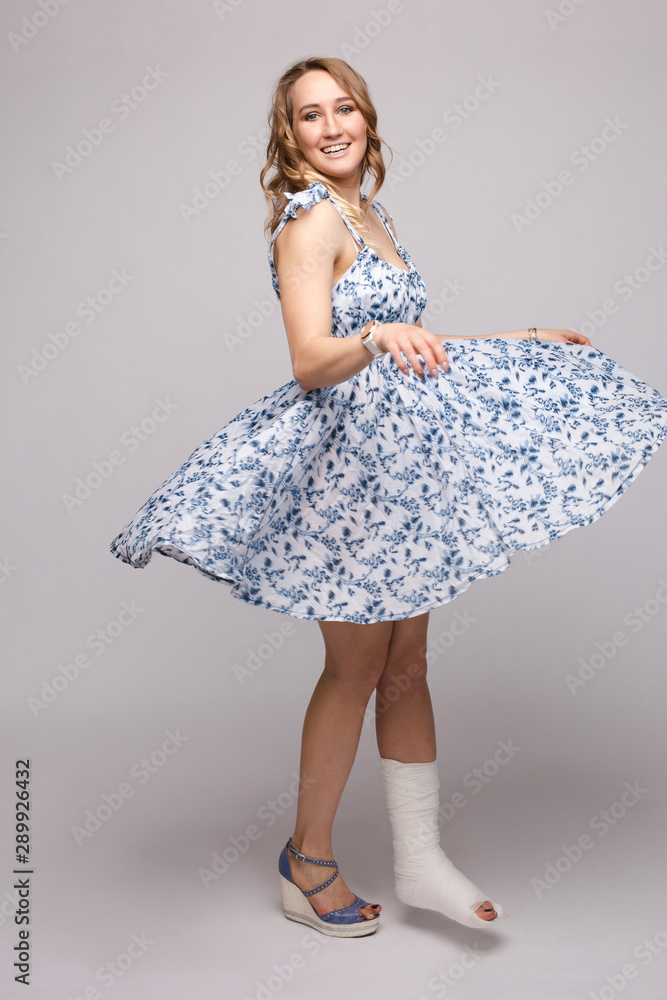 Naklejka premium Full length portrait of positive beautiful lady in summer dress and sandals dancing with her skirt flying with plaster on her broken leg. Isolate on grey. Smiling woman dancing with plaster.