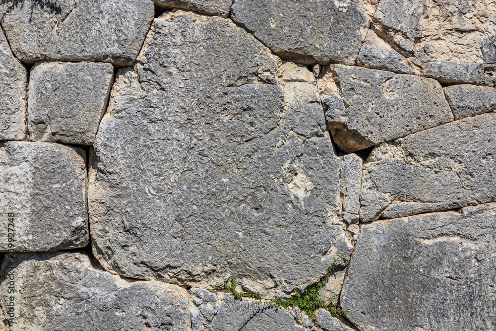 The ancient megalithic walls surround the acropolis of Amelia, in Umbria, Italy. Texture composed ofirregular huge blocks of stone. The grass grows between the interstices of the wall. The ancient megalithic walls surround the acropolis of Amelia, in Umbria, Italy. Texture composed ofirregular huge blocks of stone. The grass grows between the interstices of the wall.
