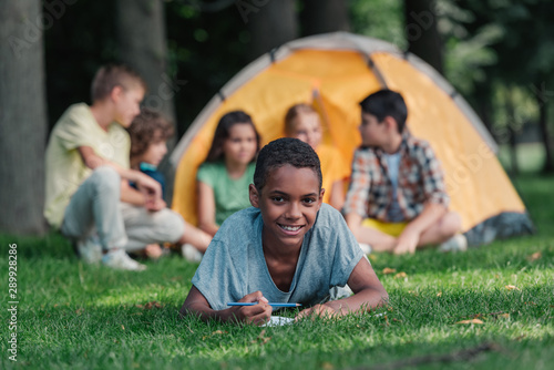selective focus of happy african american boy holding pencil near notebook and friends in camp