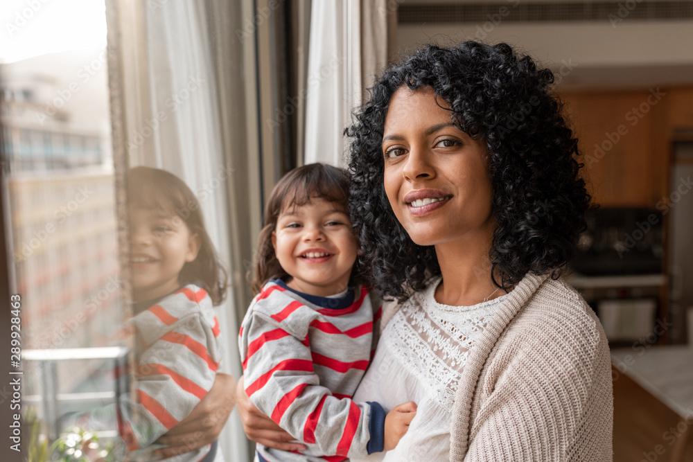 Smiling mother and daughter standing by their living room window - obrazy, fototapety, plakaty 