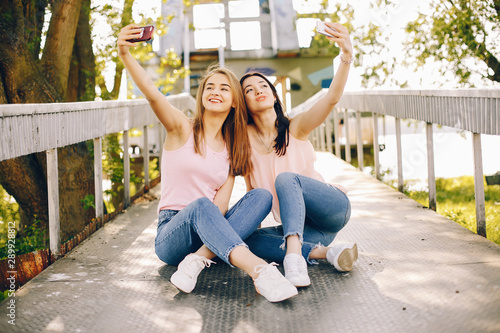 two beautiful and bright friends in pink t-shirts and blue jeans sitting in the sunny summer park and use the phone