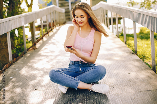 beautiful and bright girl in pink t-shirts and blue jeans sitting in the sunny summer park and use the phone with headphones