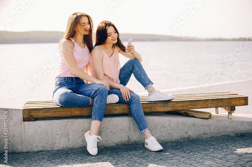 two beautiful and bright friends in pink t-shirts and blue jeans sitting in the sunny summer city and use the phone