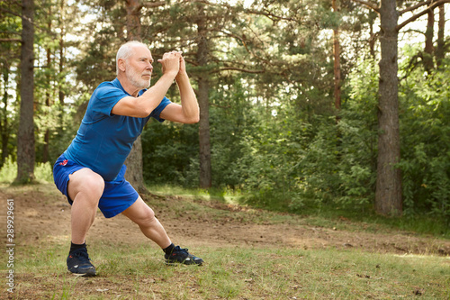 Portrait of healthy active elderly male pensioner in running shoes exercising outdoors, holding hands together in front of him and doing side lunges, having focused concentrated facial expression