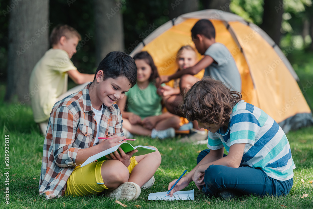 selective focus of happy boys writing on notebooks near multicultural friends in camp selective focus of happy boys writing on notebooks near multicultural friends in camp