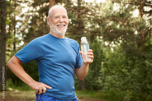 Portrait of joyful active Caucasian retired man with beard and bold head holding hand on his waist and drinking fresh water from glass bottle, having rest after morning physical workout in park