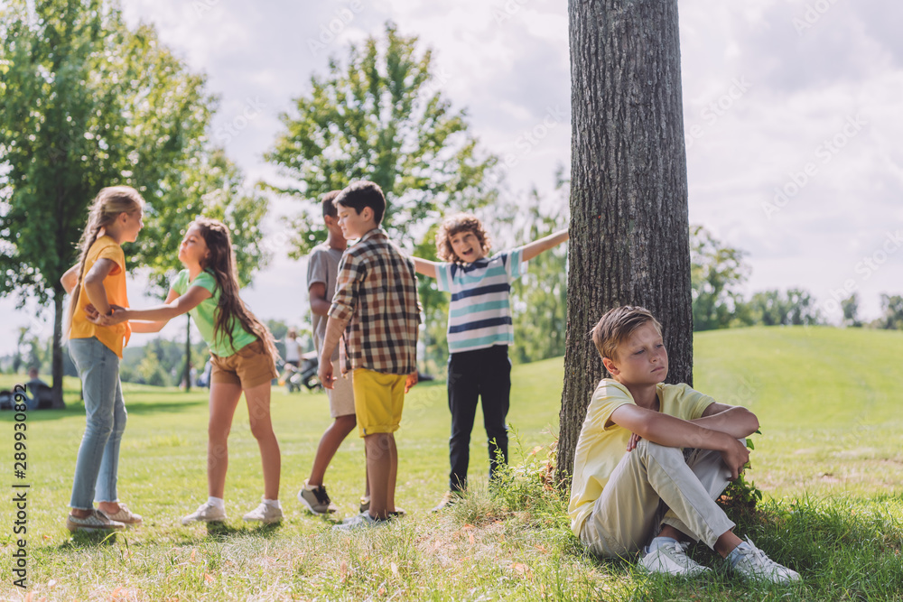 selective focus of sad boy sitting near tree trunk and multicultural children