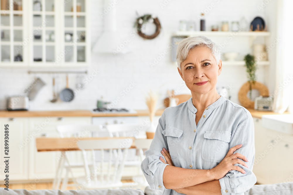 Fashionable senior female real estate agent with gray short hair standing in nice living room interior having confident facial expression, keeping arms crossed on her chest, waiting for clients Fashionable senior female real estate agent with gray short hair standing in nice living room interior having confident facial expression, keeping arms crossed on her chest, waiting for clients