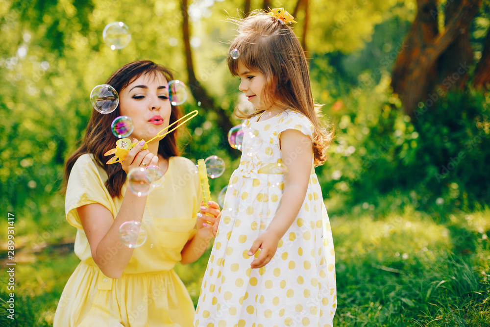 in a summer solar park near green trees, mom walks in a yellow dress and her little pretty girl and playing with bubbles in a summer solar park near green trees, mom walks in a yellow dress and her little pretty girl and playing with bubbles