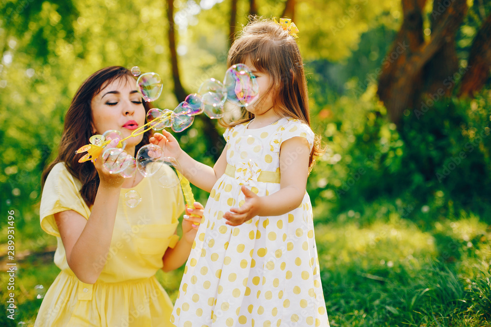 in a summer solar park near green trees, mom walks in a yellow dress and her little pretty girl and playing with bubbles