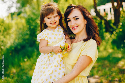in a summer solar park near green trees, mom walks in a yellow dress and her little pretty girl with yellow flowers