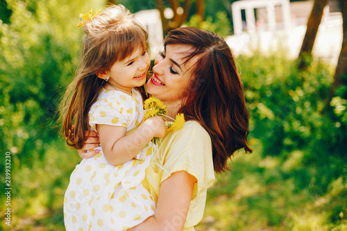 in a summer solar park near green trees  mom walks in a yellow dress and her little pretty girl with yellow flowers