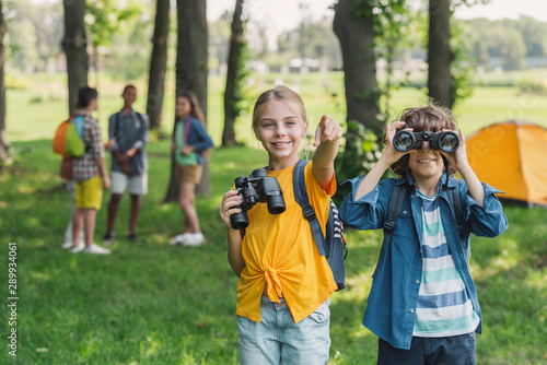 Selective focus of kid looking through binoculars near friend pointing with finger