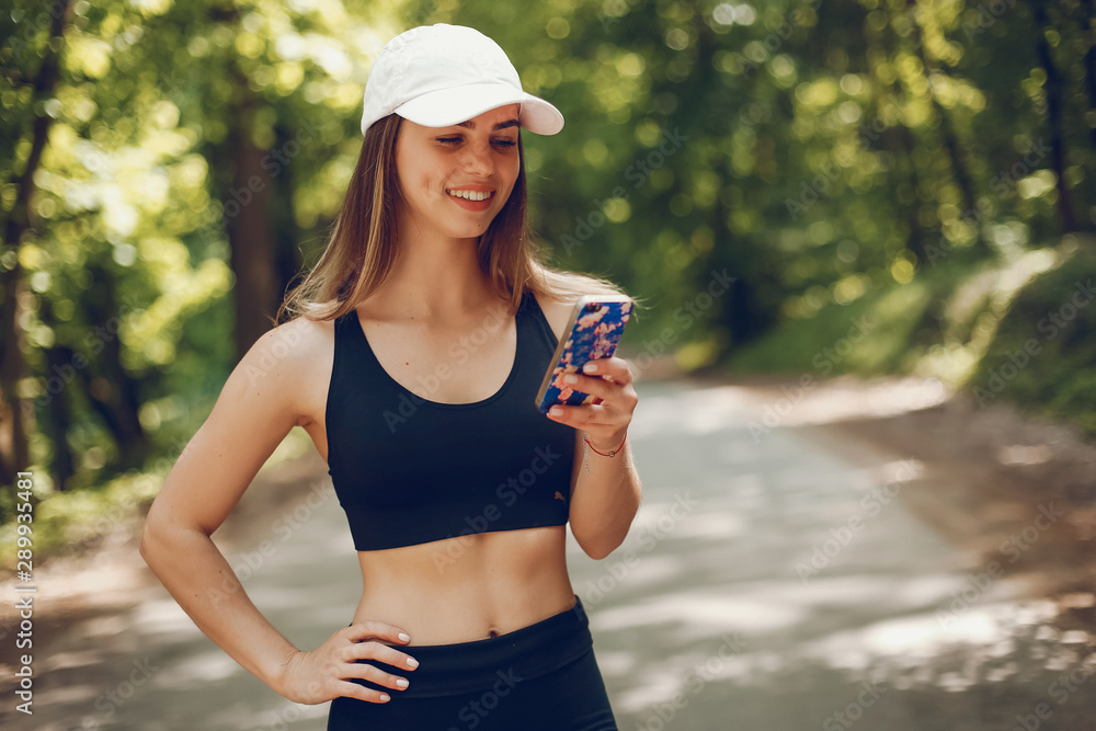 Cute girl training in a summer forest. Lady have fun in a park. Sports woman in a black sportsuit. Famale in a white cap use the phone