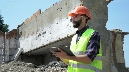 Bearded builder in a helmet and reflective vest uses a tablet on the background of a destroyed building, preparing for demolition and construction
