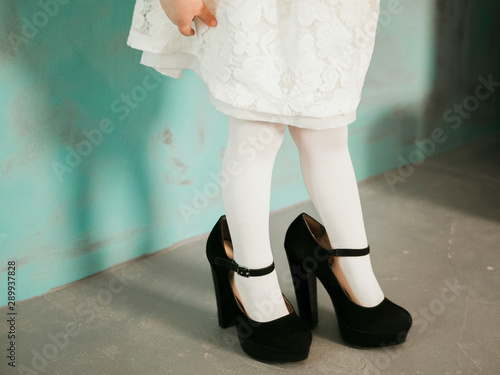 Billede på lærred Little girl in oversized black heels shoes and white dress near neo mint wall wi