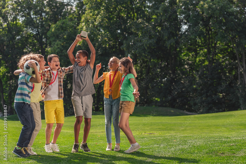 Obraz premium happy multicultural kids jumping on grass with football and trophy