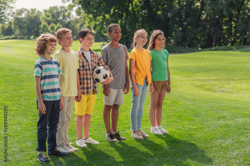 happy multicultural kids standing on grass with football