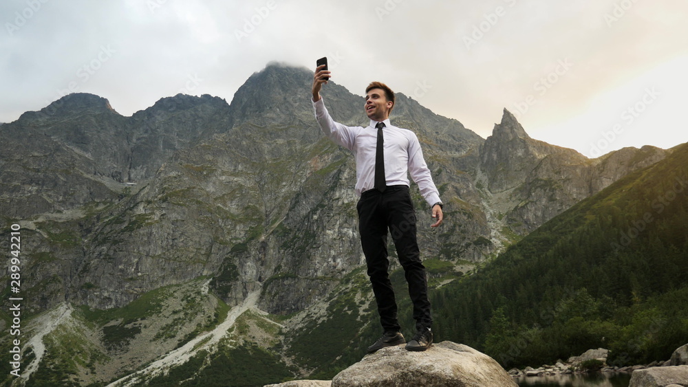 Obraz premium Successful confident businessman talking by video chat, uses a mobile phone on online conference near a lake in the rocky mountains at sunset, way to success