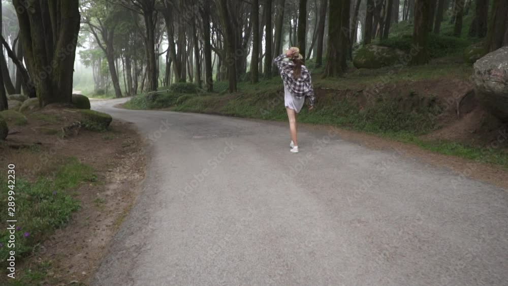 Beautiful traveler girl in dress and straw hat walking on asphalt road in magic fabulous forest with high trees and large stones. Sintra. Portugal. Beautiful traveler girl in dress and straw hat walking on asphalt road in magic fabulous forest with high trees and large stones. Sintra. Portugal.