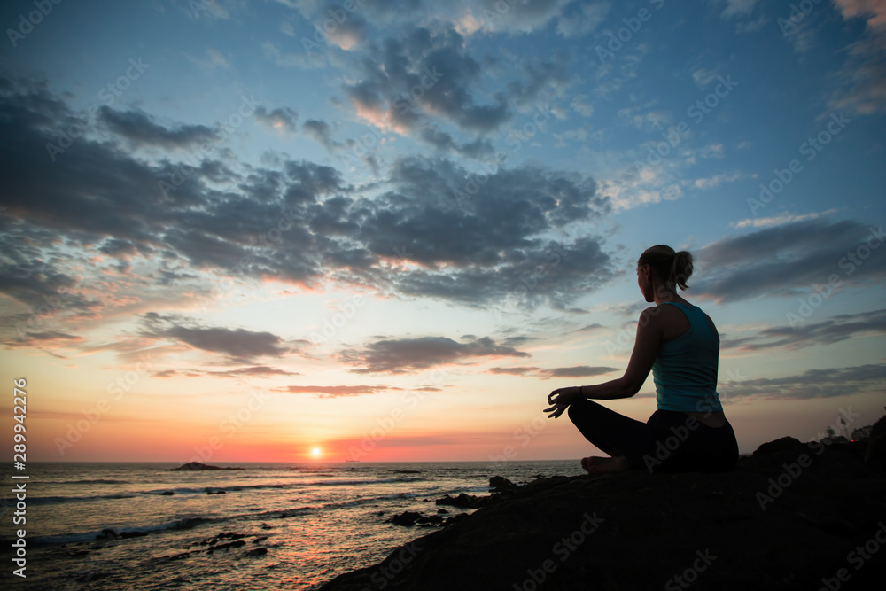 Young woman practicing yoga sitting in Lotus position on the ocean coast during sunset.