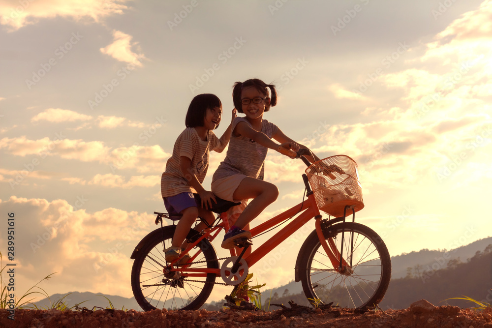 Silhouette two children playing riding bike on mountain at sunset sky background