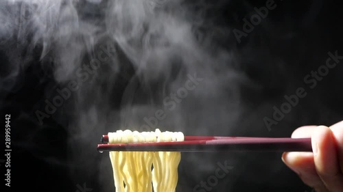 Chopsticks to tasty noodles with steam and smoke in bowl on black background in slow motion, Junk food and fast food concept, Asian meal close-up