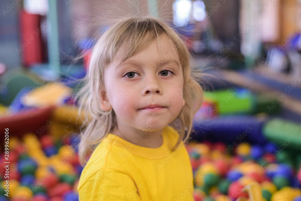 Portrait of a blond boy in a yellow t-shirt. The child smiles and plays in the children's playroom. Ball pool. Portrait of a blond boy in a yellow t-shirt. The child smiles and plays in the children's playroom. Ball pool.