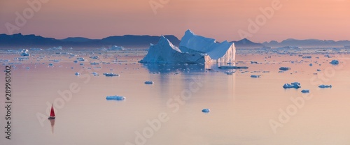 Little red sailboat cruising among floating icebergs in Disko Bay glacier during midnight sun season of polar summer. Ilulissat, Greenland. Unesco world heritage