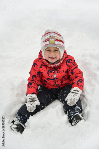 A child in a red jacket playing in the snow. The cute boy walks in the winter on the street. The kid laughs and looks at the camera. Happy childhood, fun weekend in nature. It snowed,winter is coming.