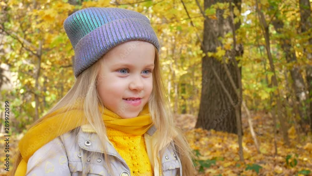 Little child in neon hat and orange scarf with finger shows something and smiles in forest or park on background of yellow foliage in autumn Little child in neon hat and orange scarf with finger shows something and smiles in forest or park on background of yellow foliage in autumn