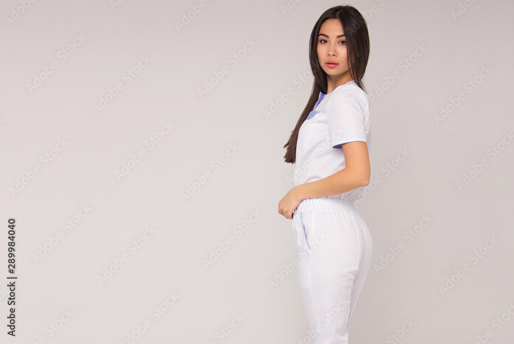 young stylish happy nurse with long brunette hair in white overalls is standing from the back, looking straight and smiling cute on white wall background. medical concept. free space on left side young stylish happy nurse with long brunette hair in white overalls is standing from the back, looking straight and smiling cute on white wall background. medical concept. free space on left side