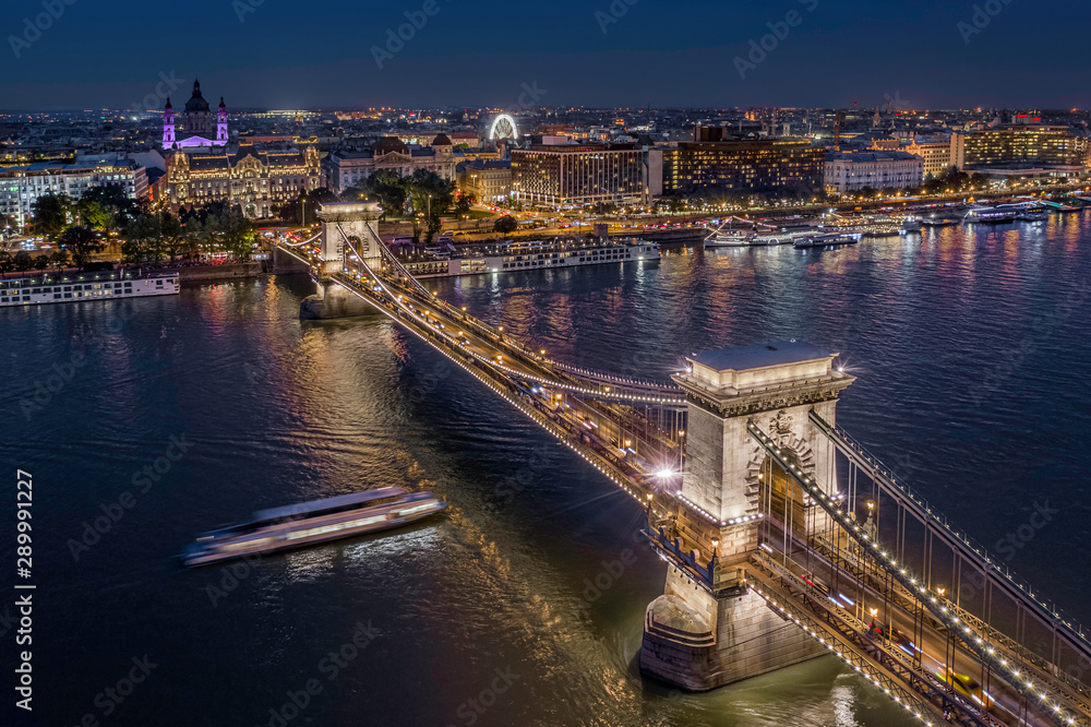 Budapest, Hungary - Aerial drone view of the beautiful illuminated Szechenyi Chain Bridge on a nice summer evening with sightseeing boat on River Danube and St. Stephens' Basilica at background