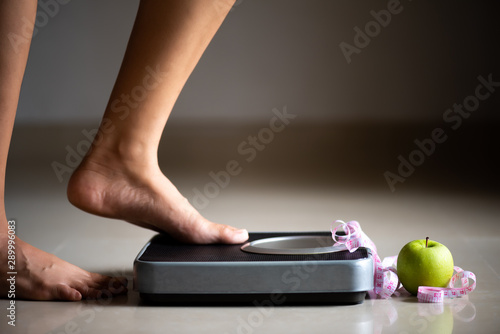 Female leg stepping on weigh scales with measuring tape and green apple. Healthy lifestyle, food and sport concept.
