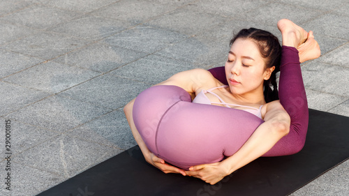 Side view of healthy women in sportswear practicing yoga outdoor. Ashtanga Yoga pose Both Legs behind Head with eyes closed, working out wearing sportswear pink top and pants, full length portrait. photo