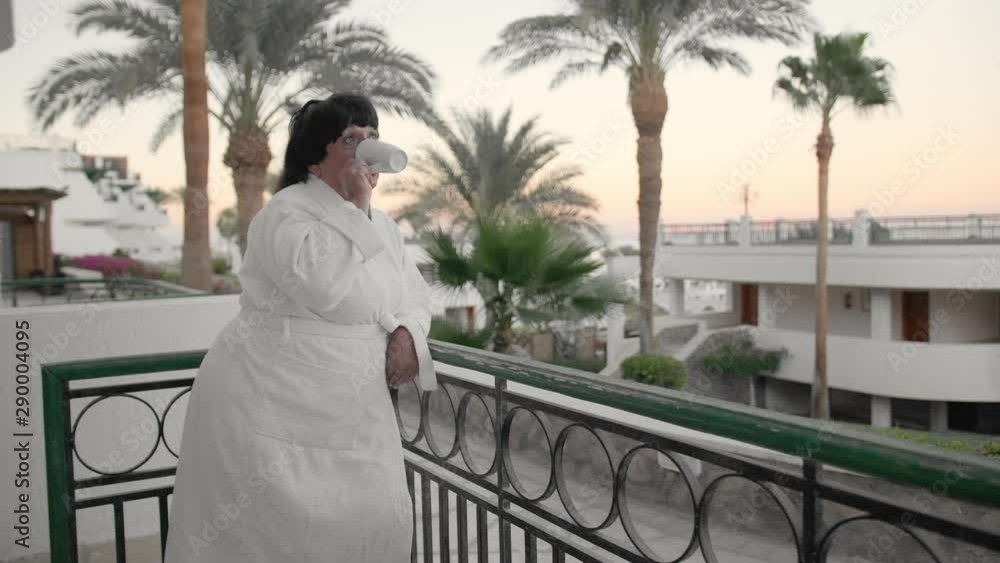 Caucasian senior woman in the morning drinking coffee on the hotel terrace in a white terry robe at a tropical resort. Against the backdrop of palm trees and the sea. Slow Motion. Caucasian senior woman in the morning drinking coffee on the hotel terrace in a white terry robe at a tropical resort. Against the backdrop of palm trees and the sea. Slow Motion.