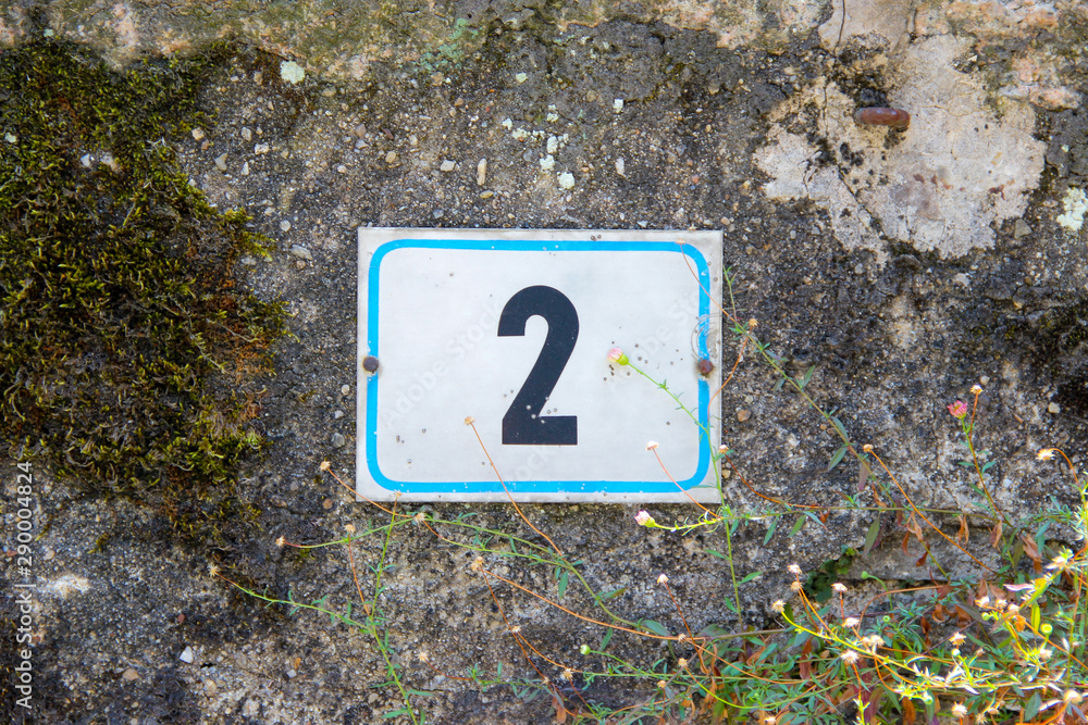 House number two. Black lettering with blue frame on a white metal plate nailed to an old damaged stone wall House number two. Black lettering with blue frame on a white metal plate nailed to an old damaged stone wall