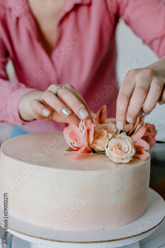 Female hand decorating pink flower wedding birthday cake on stand. Close up pastry chef decoration delicious dessert dish.