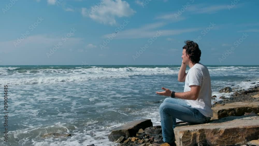 Upset young man swears with someone on the phone sitting on the beach. Throws phone into the sea Upset young man swears with someone on the phone sitting on the beach. Throws phone into the sea
