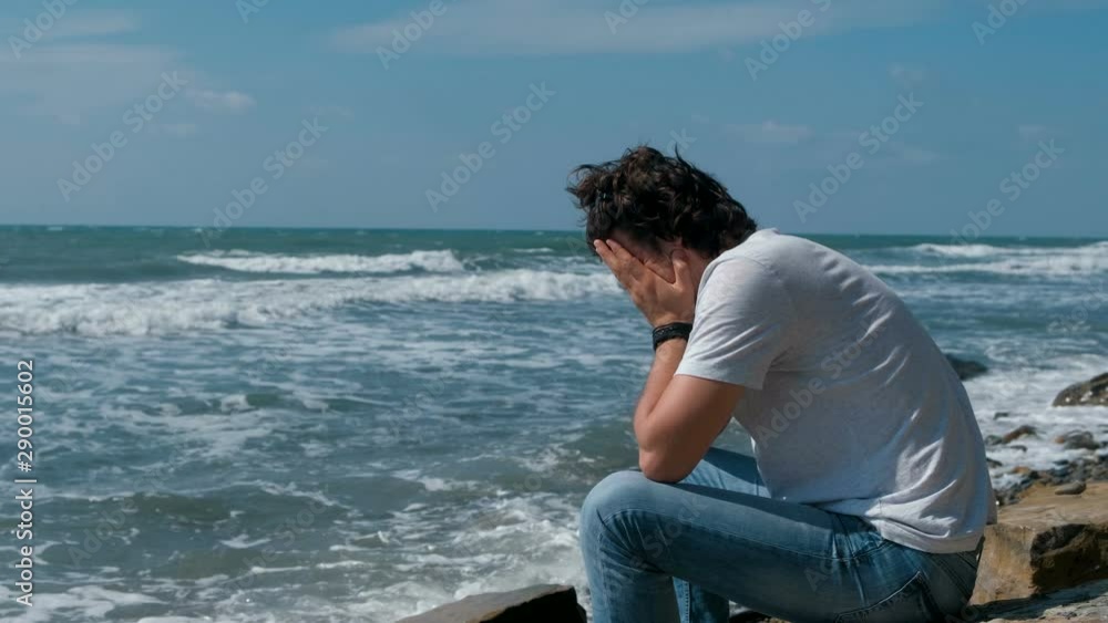 Upset young man sitting on the beach and looking at waves Upset young man sitting on the beach and looking at waves